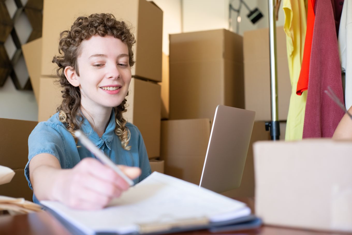 Small business owner writing notes at a desk with a laptop, surrounded by cardboard boxes