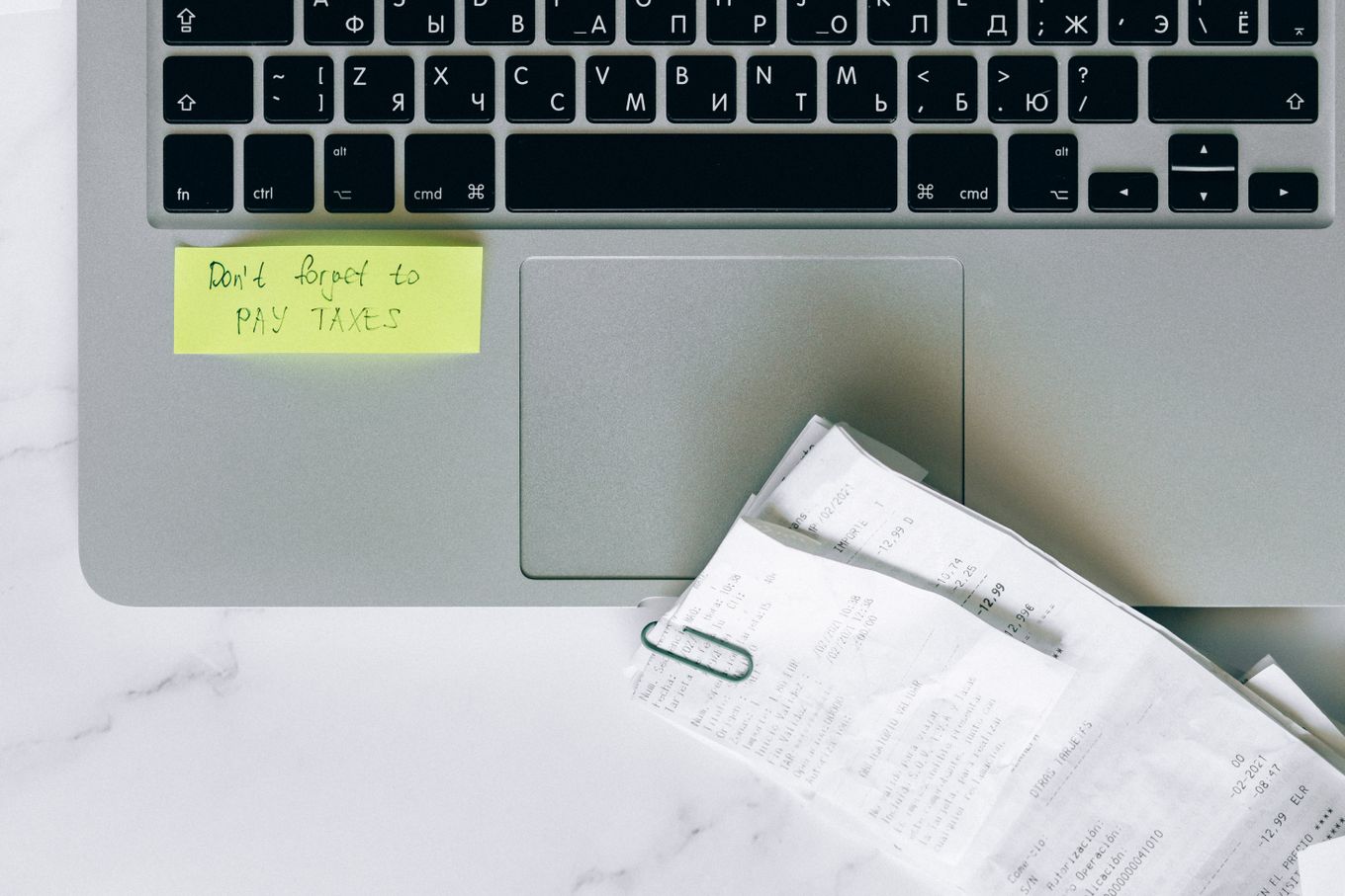 Laptop with a sticky note reading don't forget to pay taxes next to a stack of receipts