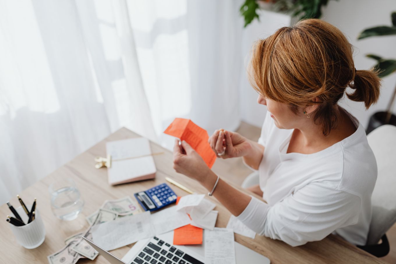 Woman at a home office desk sorting receipts with a calculator and laptop