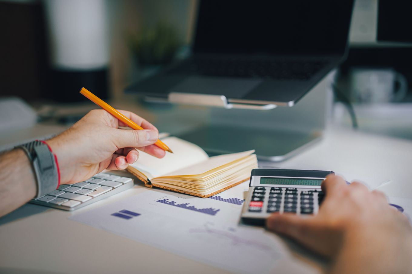 Hands using a calculator and taking notes at a desk with a laptop and printed charts